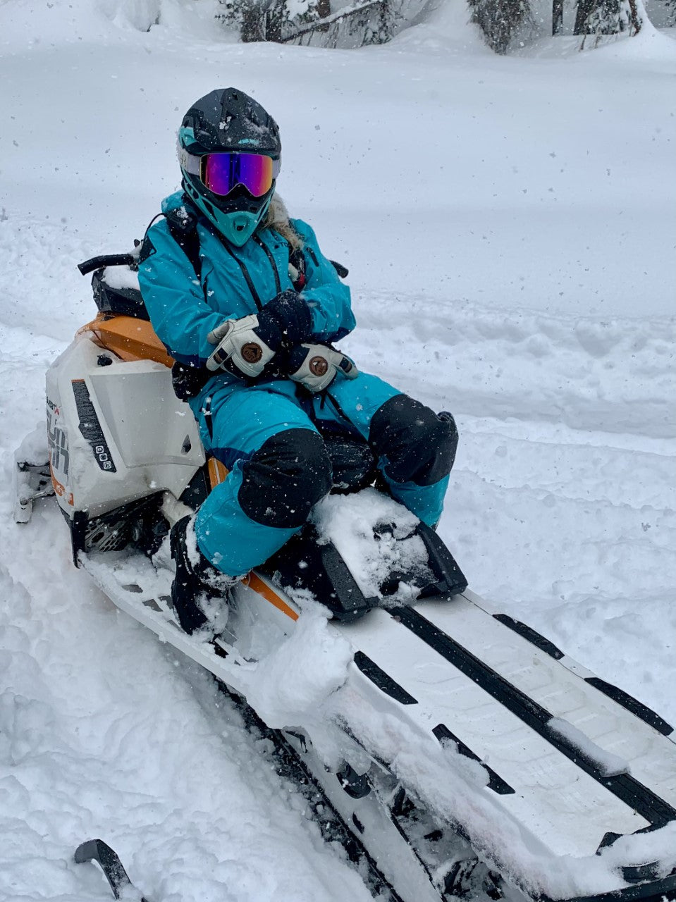 Person seated on a snowmobile against a snowy backdrop, dressed in blue winter clothing and wearing Baist ski gloves, ready for an adventure in the cold with style and comfort.
