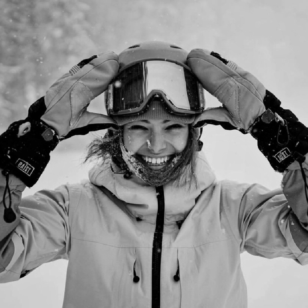 man wearing a ski jacket and BAIST mitt holding the goggles smiling with a snow covered mountains at the back