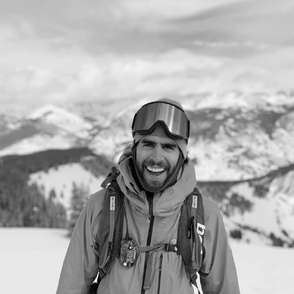 man wearing a ski jacket smiling with a snow covered mountains at the back