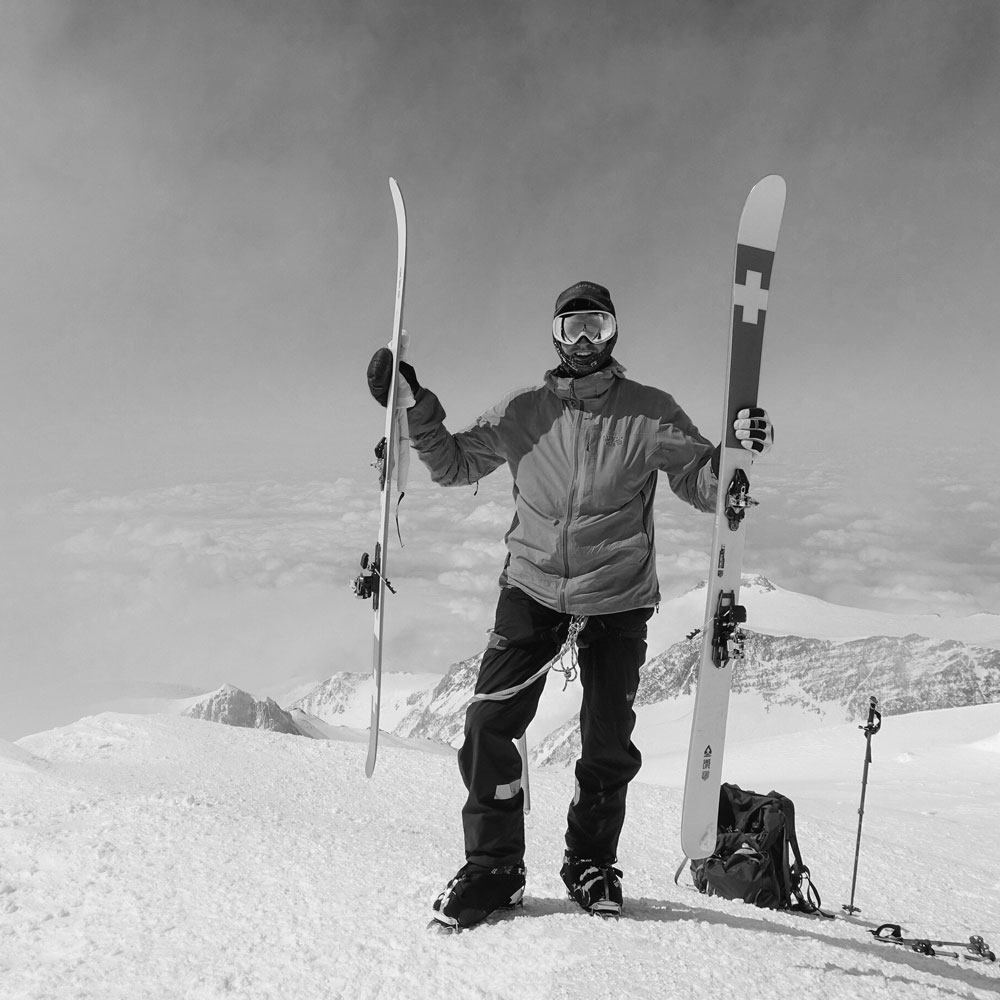 man standing on a snow holding a ski board with a snow covered mountains at the back