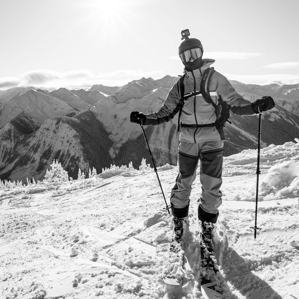 Man standing on the snow holding a ski pole wearing BAïST mitt with a snow covered mountains at the back